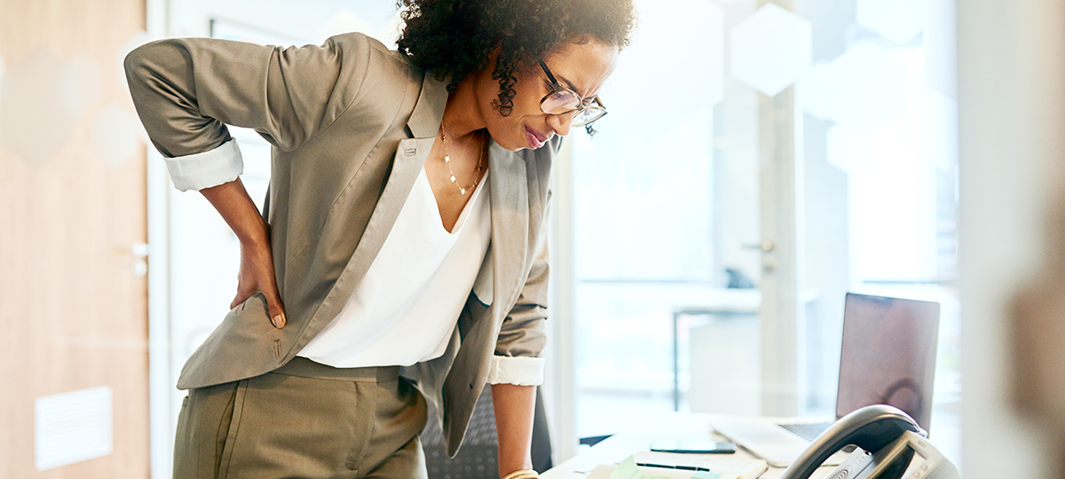 Person leaning over desk with hand on back and look of pain on face